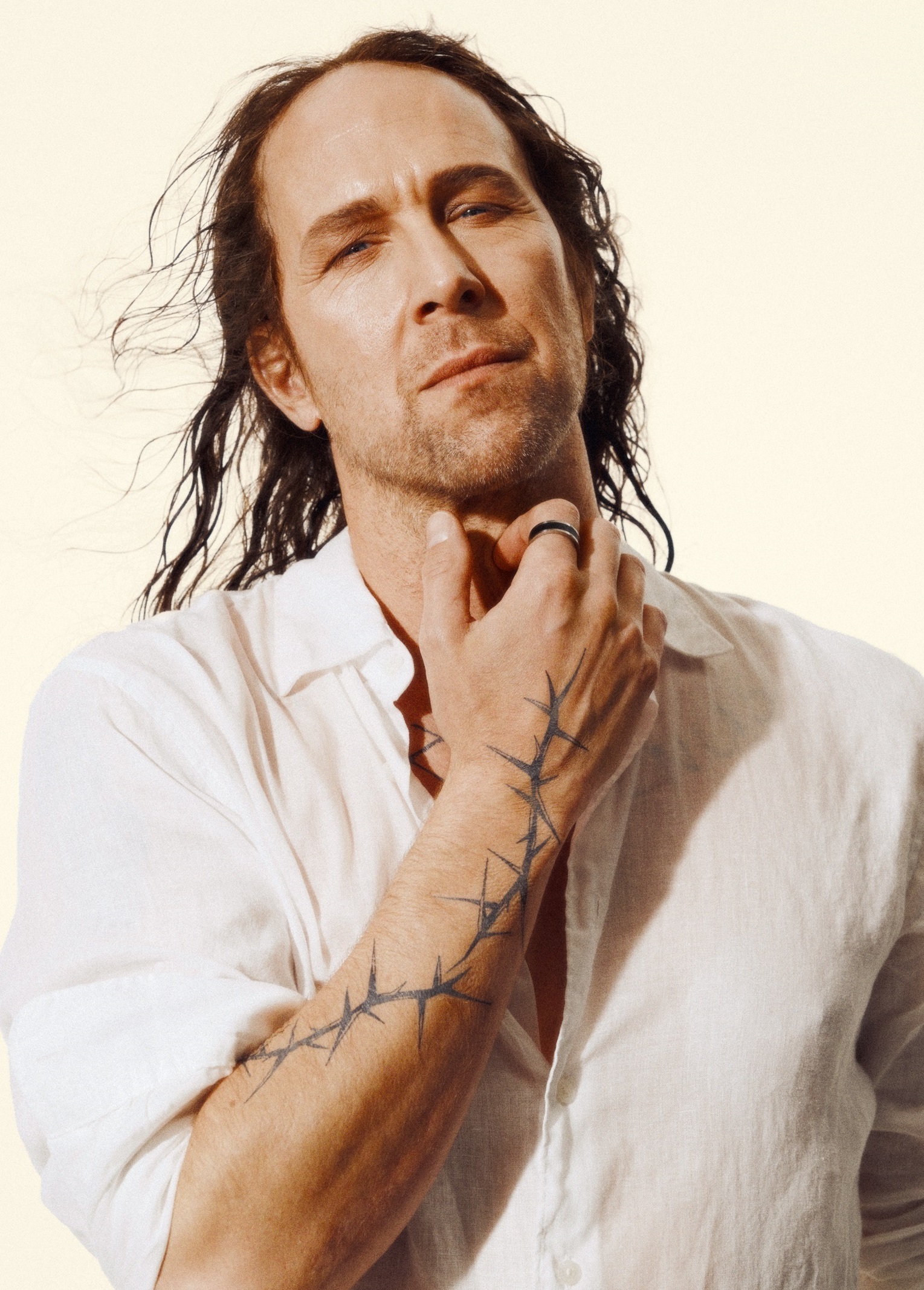 Man i 40-årsåldern med långt mörkt hår, vit skjorta och taggtrådsliknande tatuering på höger arm.