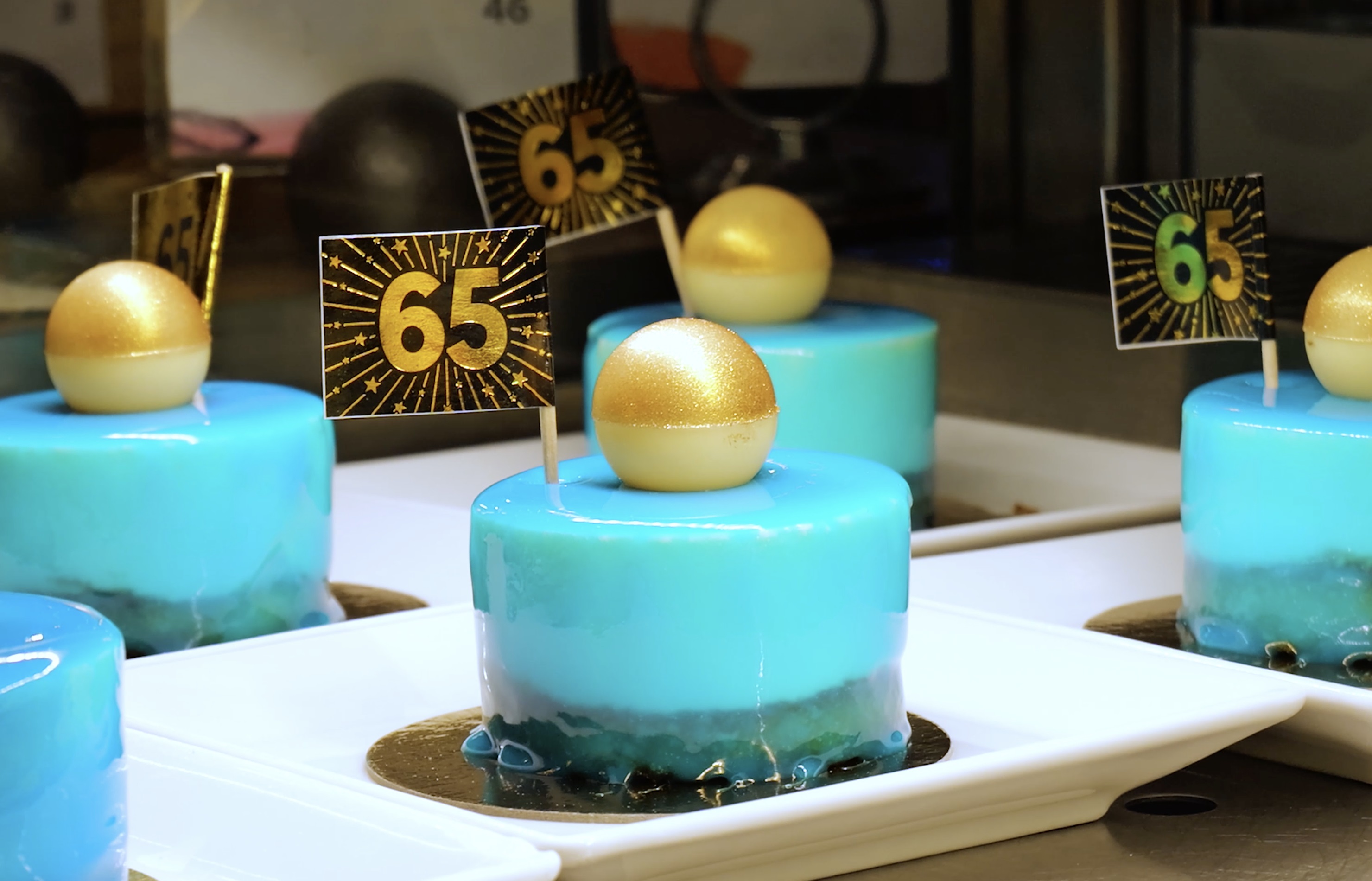 Blå glasstårtor med guldfärgade kulor och skyltar med siffran 65 på toppen, placerade på vita fat.
