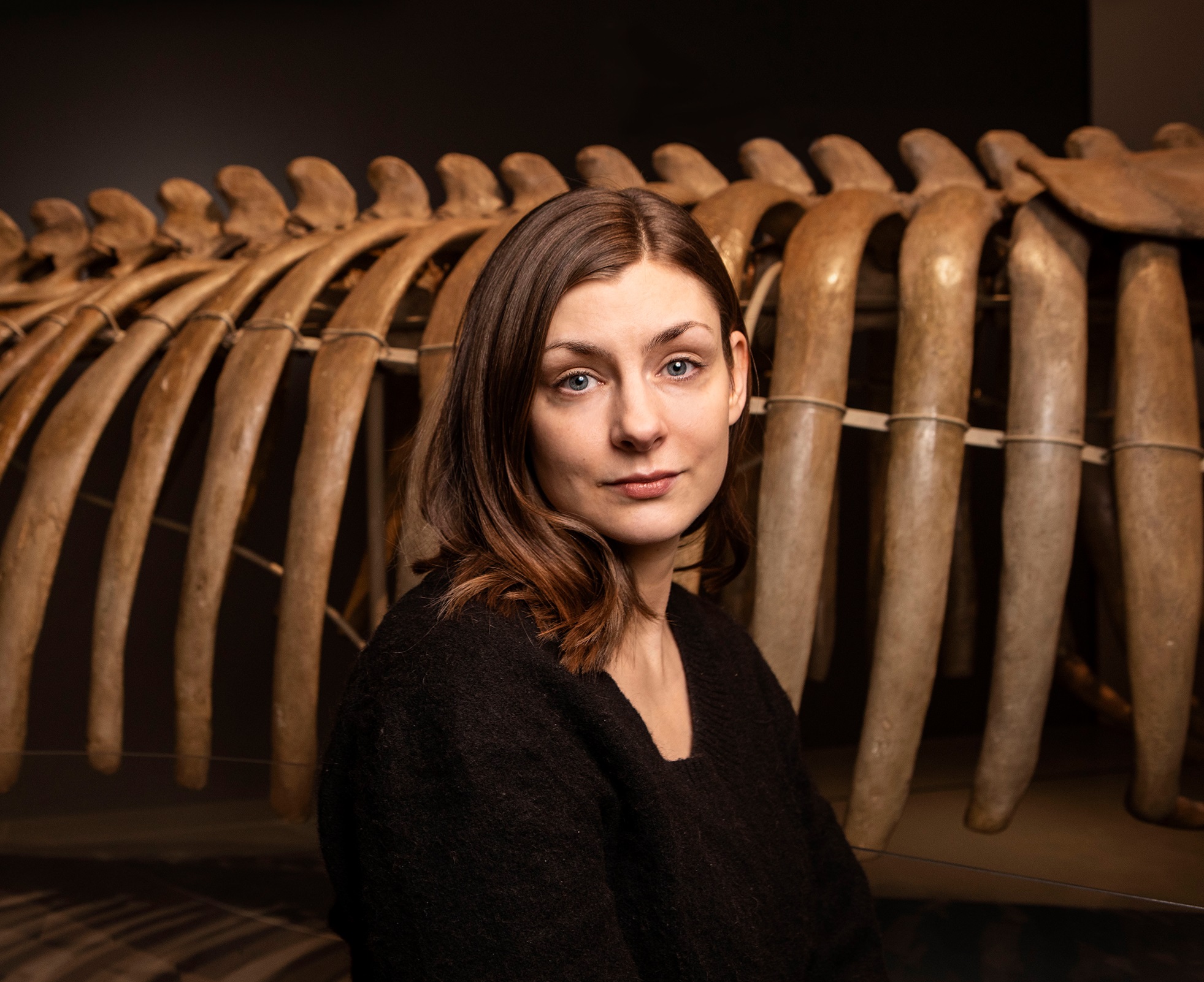 Kvinna i 30-årsåldern med brunt axellångt hår och svart tröja, står framför valskelett.