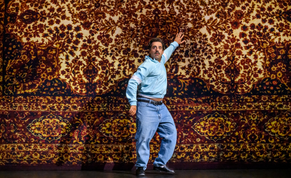 Man i blå träningsjacka och jeans poserar mot en vägg med orientaliskt mattmönster.