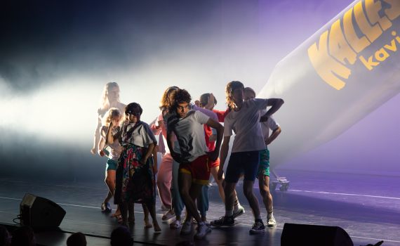 Grupp personer i scenkläder dansar framför en stor Kalles kaviar-rekvisita på scen.