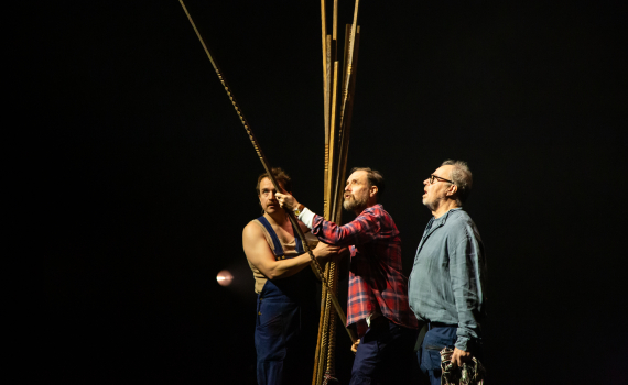 Tre män håller upp långa trästavar på mörk teaterscen, en bär flanellskjorta, en har blå snickarbyxor.