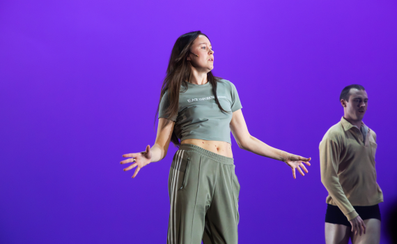 Kvinna i grönt träningsset och man i beige tröja uppträder på scen med lila bakgrund.