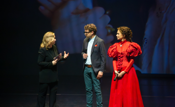 Två kvinnor och en man står på scen, en kvinna bär röd klänning i äldre stil, de andra är formellt klädda.