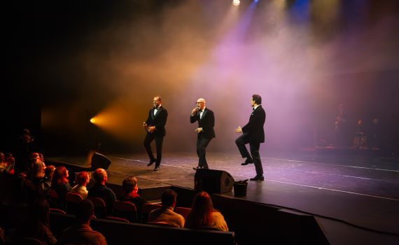 Tre män i svarta kostymer uppträder i sång och dans på scen inför sittande publik.