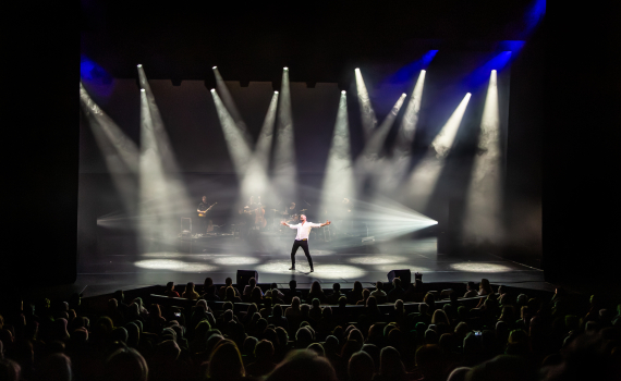 Man i vit skjorta uppträder ensam mitt på scen, upplyst av flera starka scenlampor inför publik.