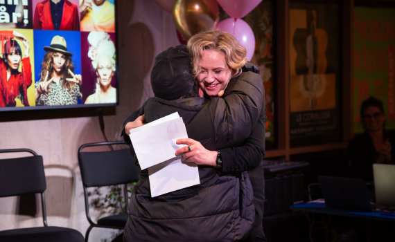 Kvinna med blont hår kramar person i svart jacka, håller papper, framför ballonger och affisch.