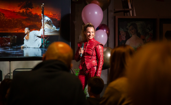 Kvinna i röd scenkostym talar i mikrofon inför publik, bakom syns affisch med teaterbild.