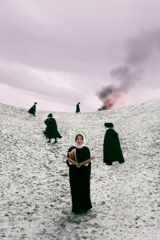 Kvinna i svart klänning och vit hätta håller bok i snötäckt landskap med flera personer och rök i bakgrunden.