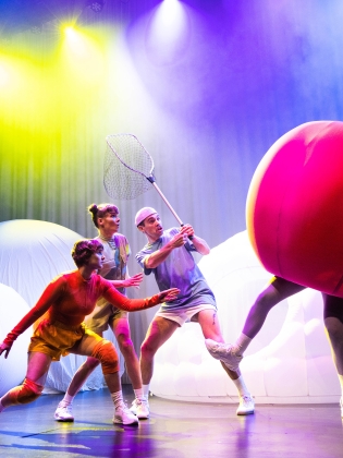 Tre personer i färgglada kläder försöker fånga in en stor rosa boll med ben i en hov. Bollen försöker fly och personerna tittar nyfiket efter den.