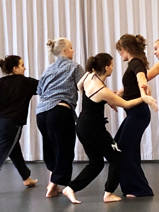 Fem unga kvinnor i träningskläder dansar barfota i en grupp framför ett vitt draperi.