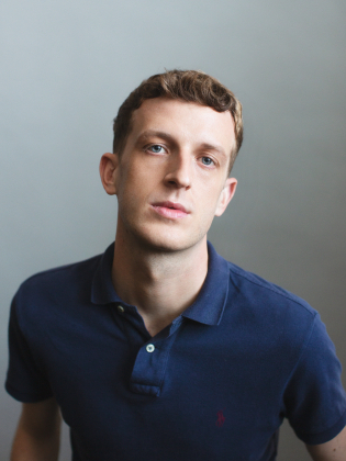 Ung man med kort lockigt hår i mörkblå pikétröja mot ljusgrå bakgrund.