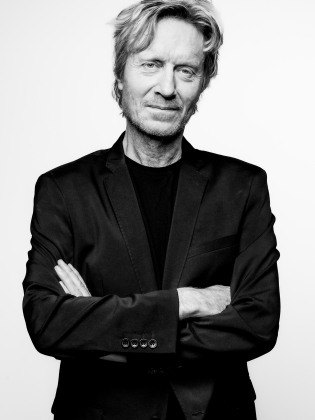 Man i 60-årsåldern med ljust, rufsigt hår och skäggstubb i svart kavaj mot vit bakgrund.