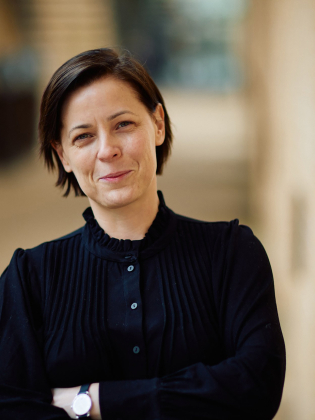 Kvinna i 30-årsåldern med kort mörkt hår och svart blus står med armarna i kors mot suddig bakgrund.