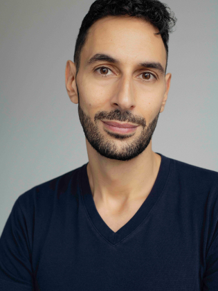 Man i 30–40-årsåldern med kort lockigt hår och skägg, i mörkblå t-shirt mot ljus bakgrund.