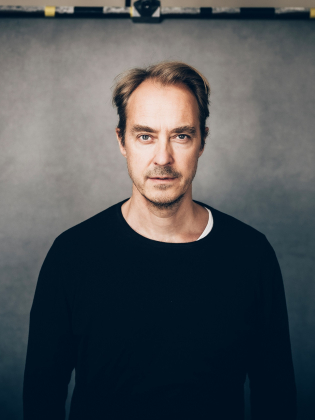 Man i 40–50-årsåldern med tunnat hår och skäggstubb, i svart tröja framför grå bakgrund.