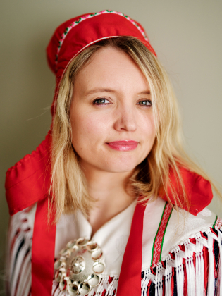 Kvinna i traditionell samisk dräkt med röd kolt och silverbrosch, blondt hår och röd huvudbonad.