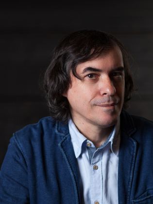 Man i medelåldern med mörkt, lockigt hår, blå skjorta och blå kavaj, mot mörk bakgrund