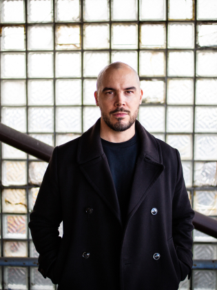 Skallig man i 30–40-årsåldern med skägg i mörk rock, framför glasblocksvägg med metallräcke.