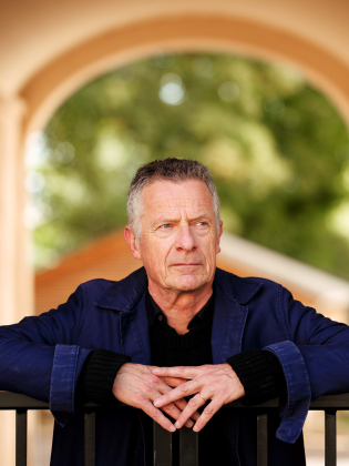 Äldre man med kort grått hår i blå jacka, lutar sig mot svart räcke i valv med suddig bakgrund.