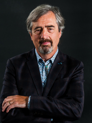 Äldre man med grått hår och skägg, bär mörk kavaj och mönstrad skjorta mot svart bakgrund.