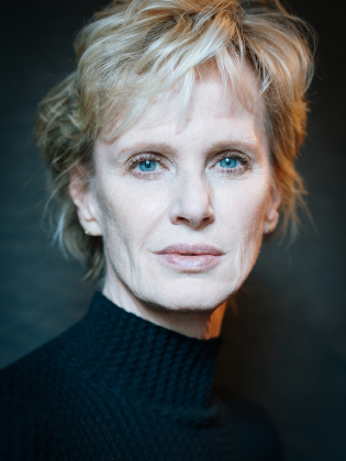 Äldre kvinna med kort blont hår och blå ögon, bär svart tröja mot mörk suddig bakgrund.