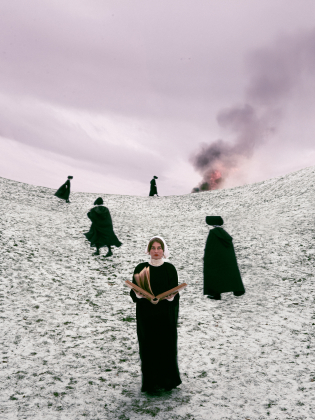 Kvinna i svart klänning och vit hätta håller bok i snötäckt landskap med flera personer och rök i bakgrunden.