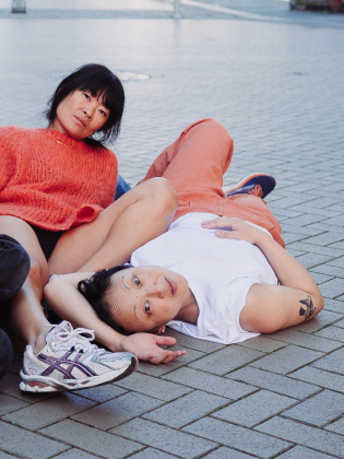 Två vuxna personer ligger på marken; en i orange tröja och en i vit t-shirt med tatuering på överarmen.