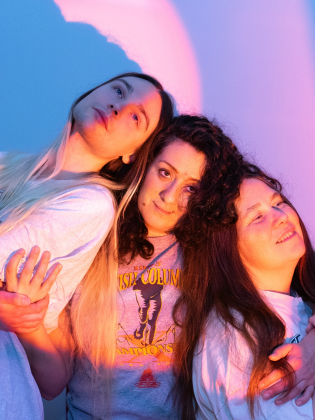 Tre unga kvinnor med långt hår står tätt ihop framför en blå vägg med rosa och lila ljus.