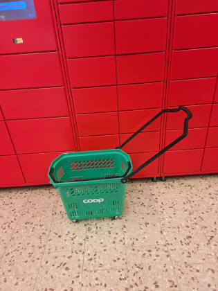 Grön rullkorg med handtag från Coop står framför röda postboxar inomhus.