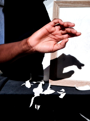 En hand i luften kastar en skuggfigur i en tom träram mot vit bakgrund med pappersbitar på bordet.