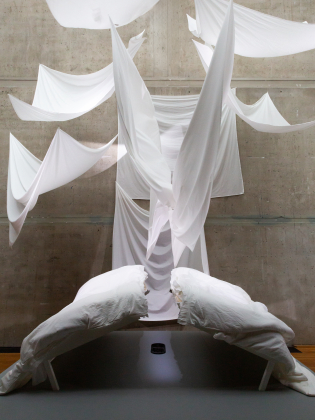 Installationskonst med upphängda vita tygstycken i olika former mot en betongvägg och två böjda sängar i förgrunden.