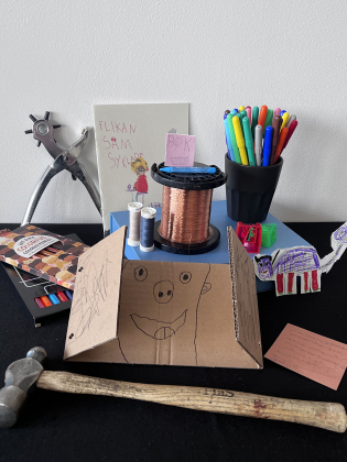 Samling av pyssel och verktyg: kartongfigur, hammare, pennor, trådrulle och barnteckningar på bord.