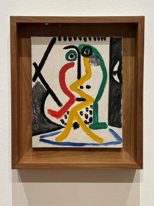 Målning i träram med abstrakt figur i gult, rött och grönt, svarta former och blå linje nederst mot vit bakgrund.