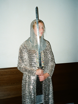 Person i glittrande rustning och ringbrynja står med ett stort svärd framför kroppen och ser åt sidan mot en neutral vägg.