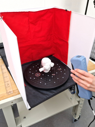 Person skannar en liten skulptur med en handhållen 3D-skanner på en roterande platta.