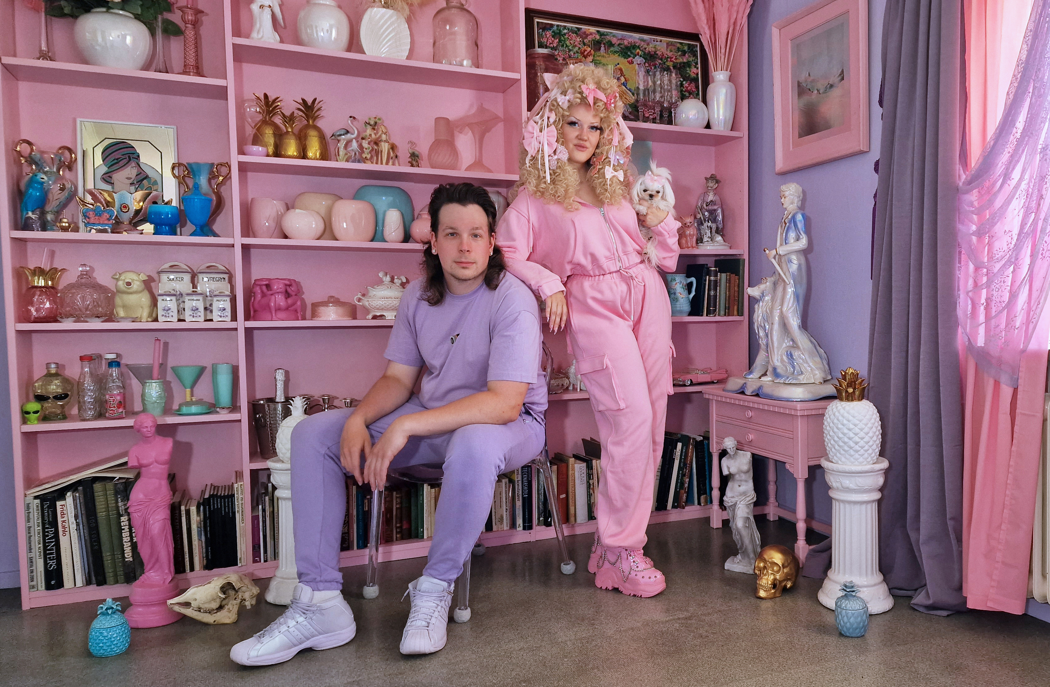 Två personer klädda i rosa och lila färger, i ett rum med rosa och lila möbler, väggar och gardiner.
