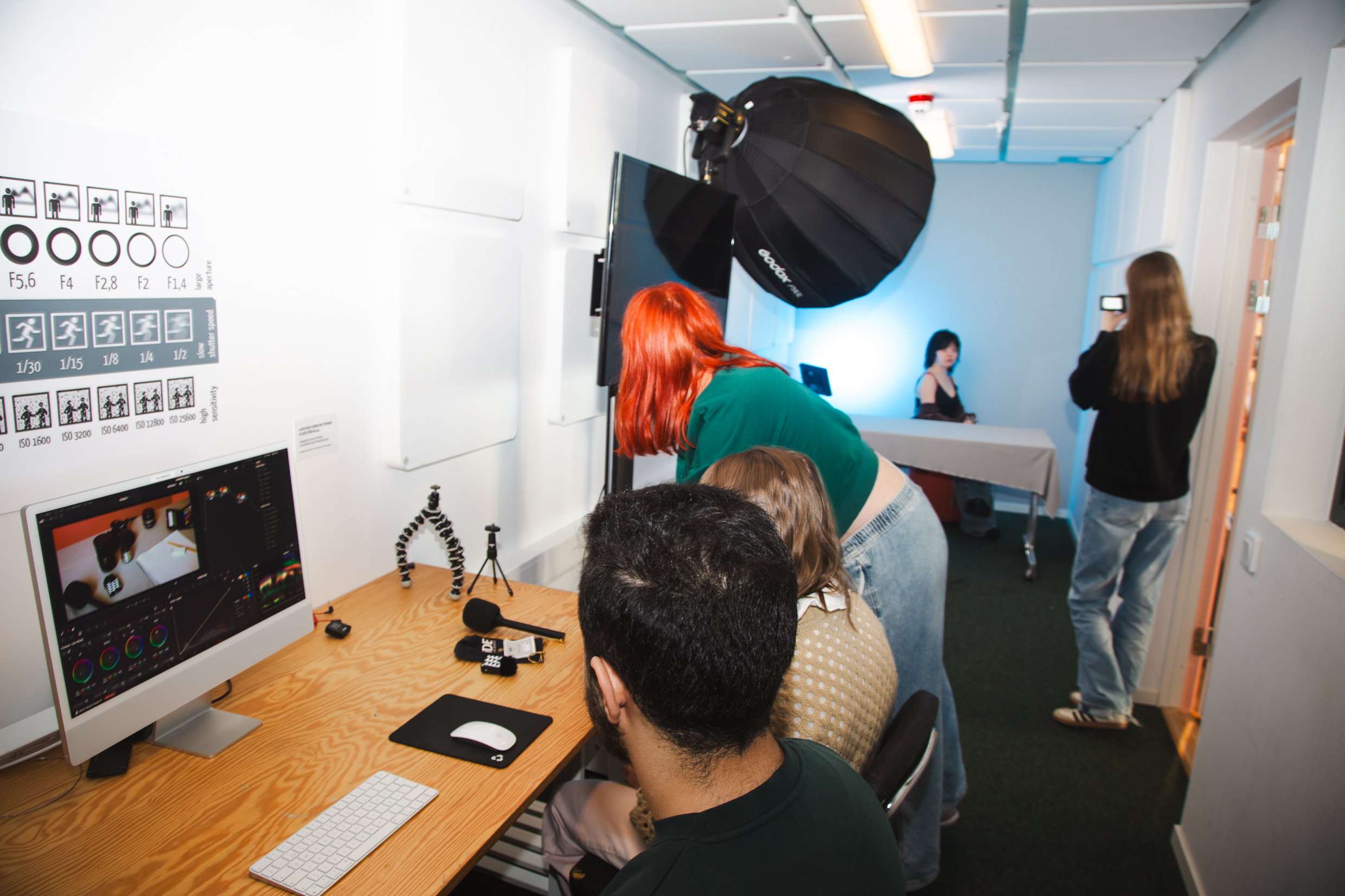 Flera personer i en studio med dator, fotoutrustning och lampor; en person fotograferar i bakgrunden.