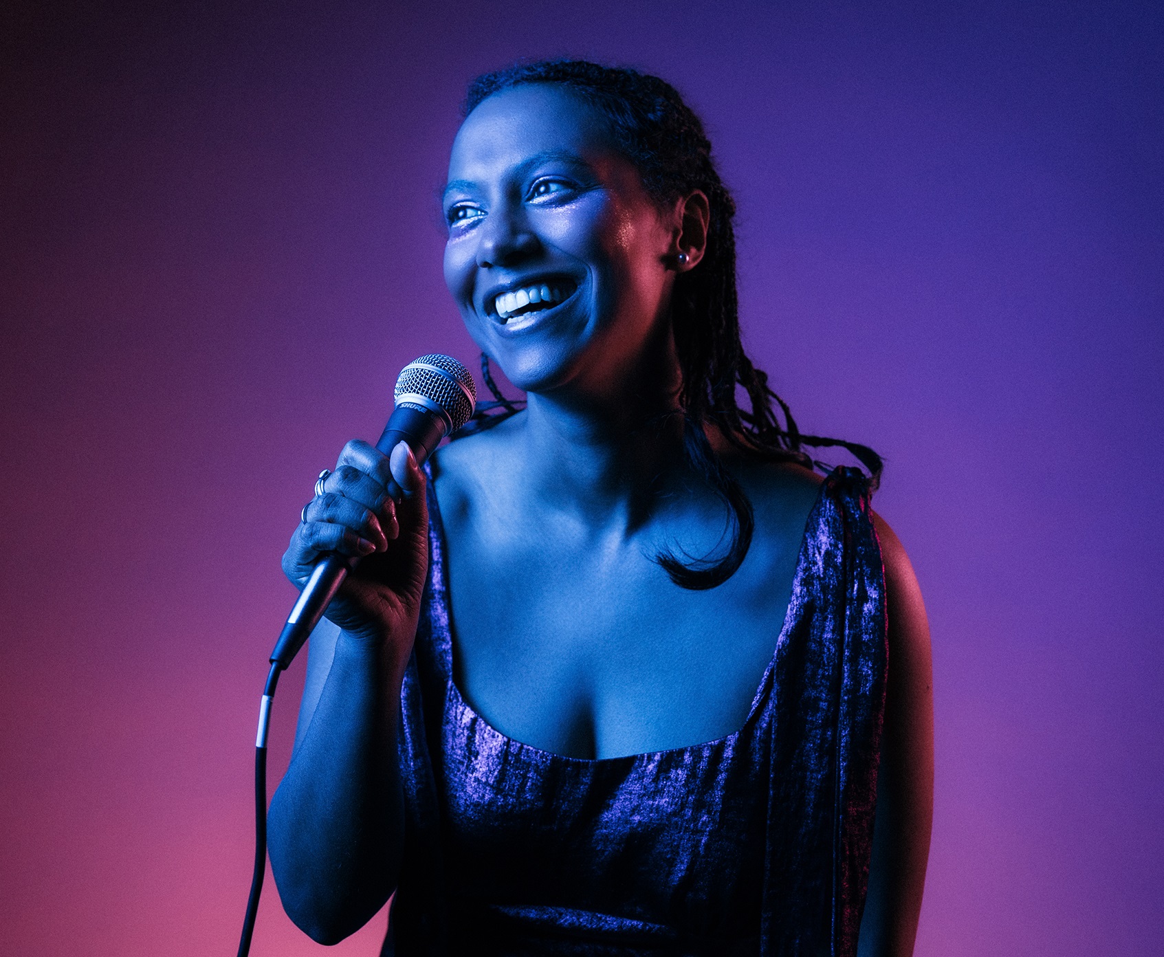 Foto: Darren Gill Kvinna med flätat hår i glittrig klänning håller mikrofon och ler mot sidan i färgat studioljussken.