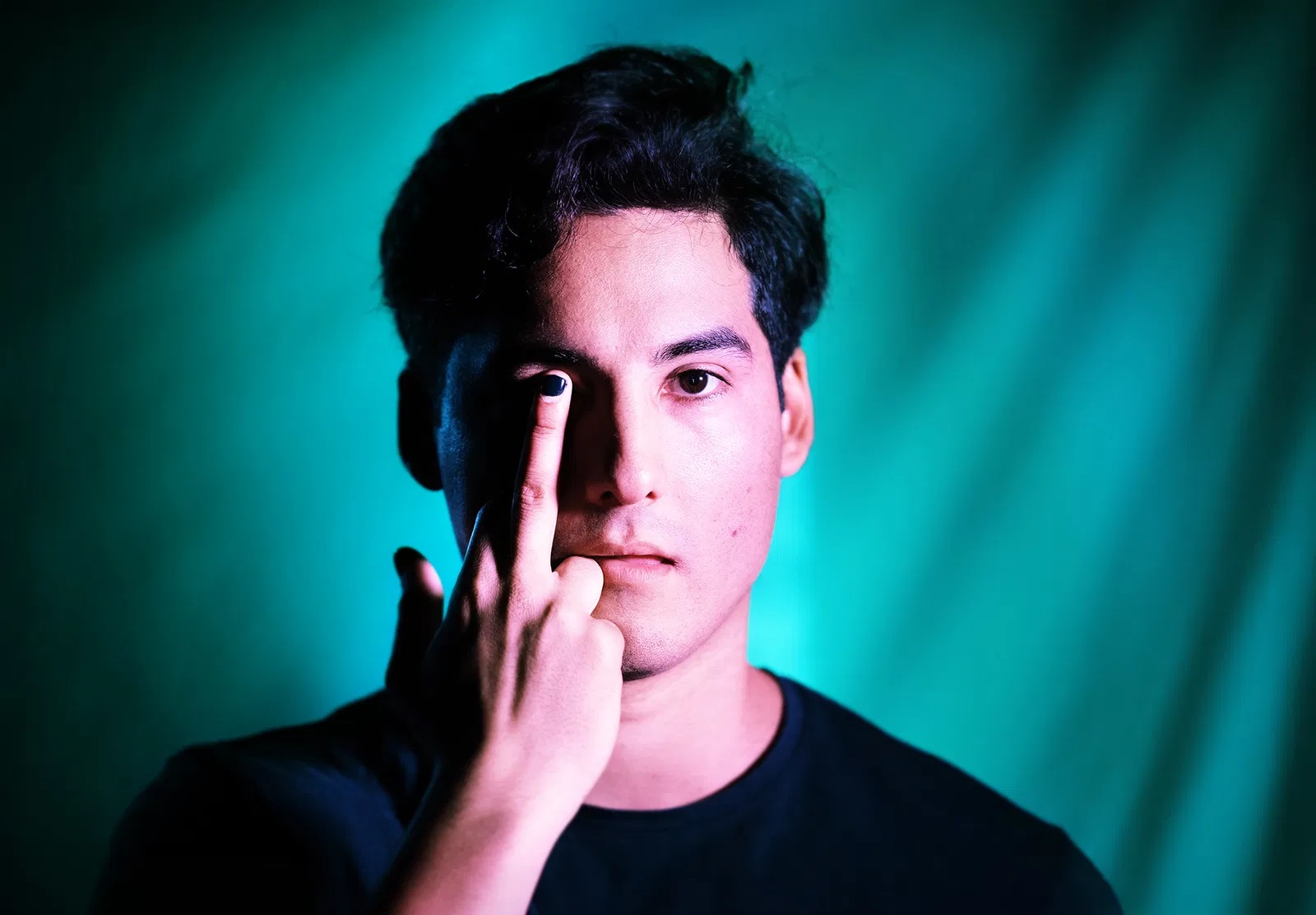 Ung man med mörkt hår håller upp ett finger med svart nagellack mot sitt öga mot turkos bakgrund.