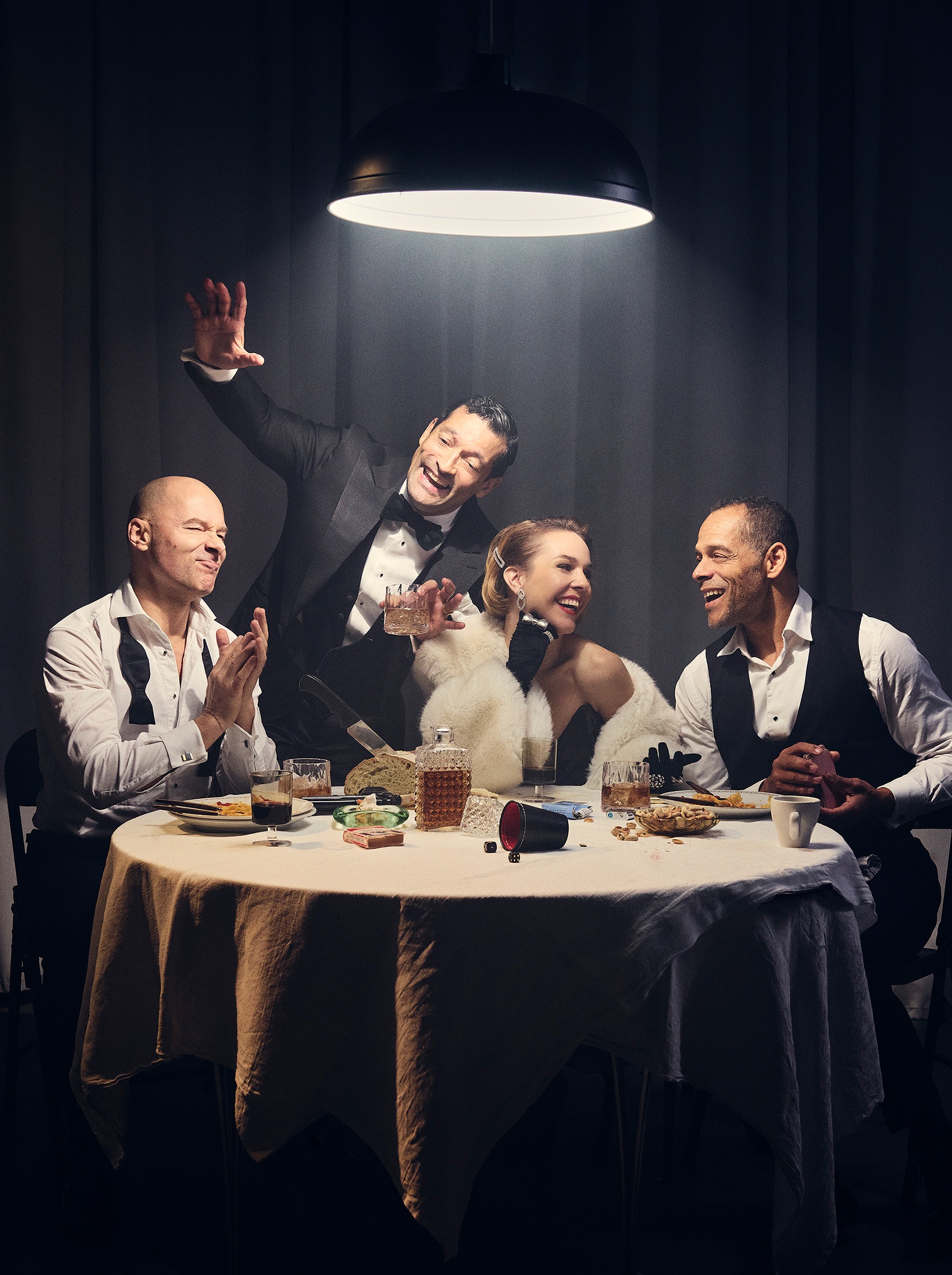 Fyra vuxna personer i festkläder runt ett bord med mat och dryck, en man står upp och gestikulerar.