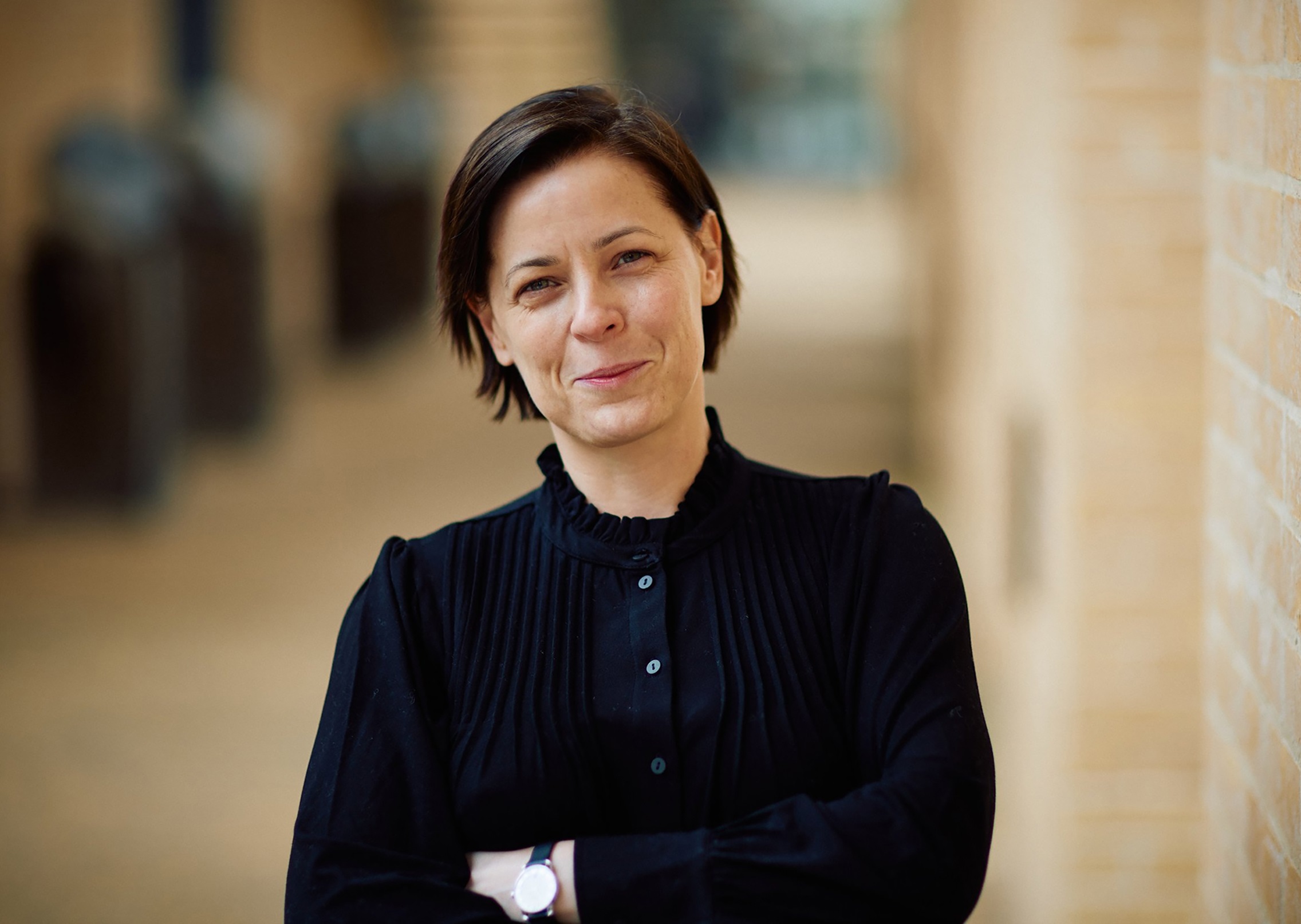 Kvinna i 30-årsåldern med kort mörkt hår och svart blus står med armarna i kors mot suddig bakgrund.