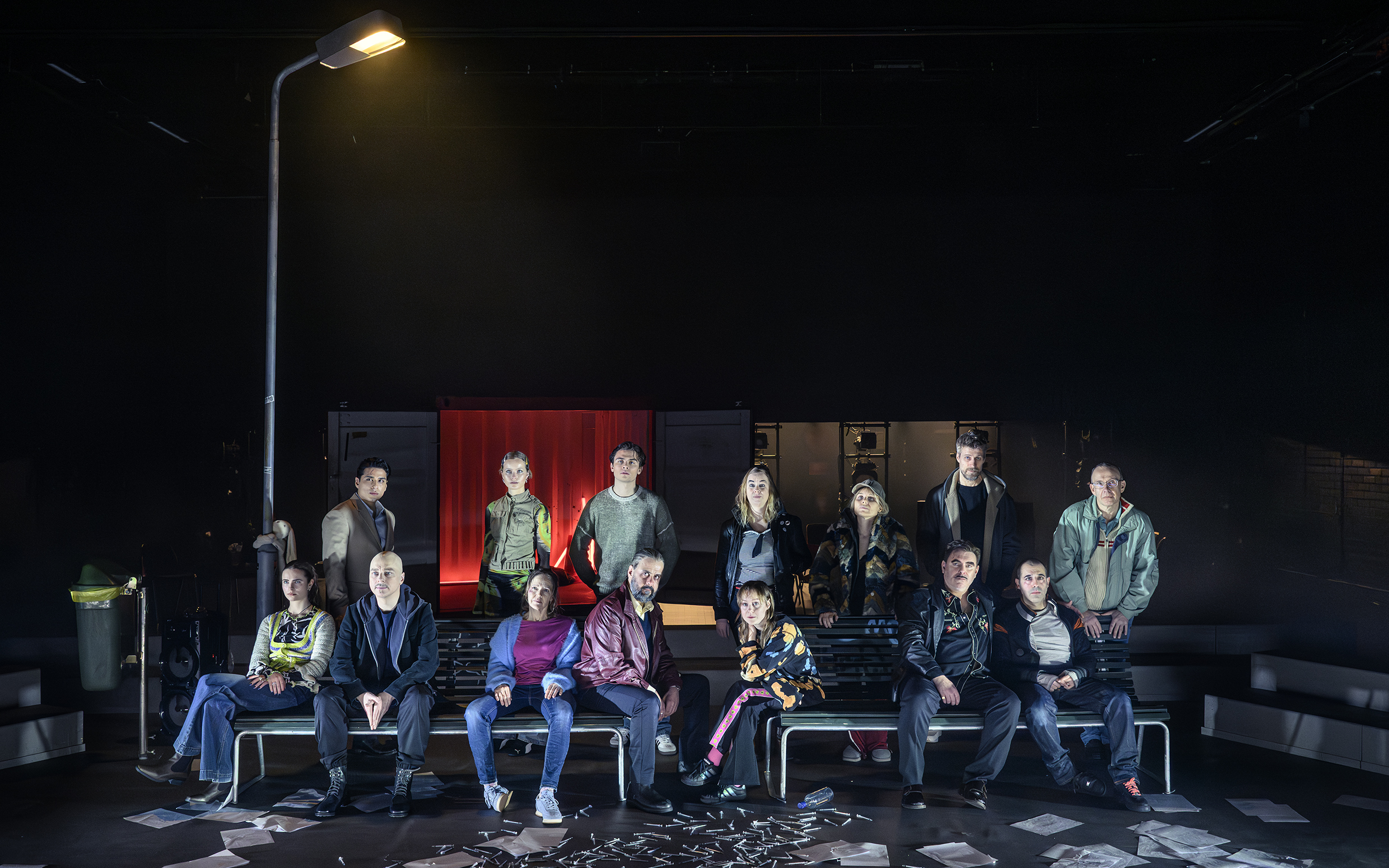 Tretton vuxna män och kvinnor i olika åldrar sitter och står på bänkar under gatlykta; sprutor och papper på golvet.