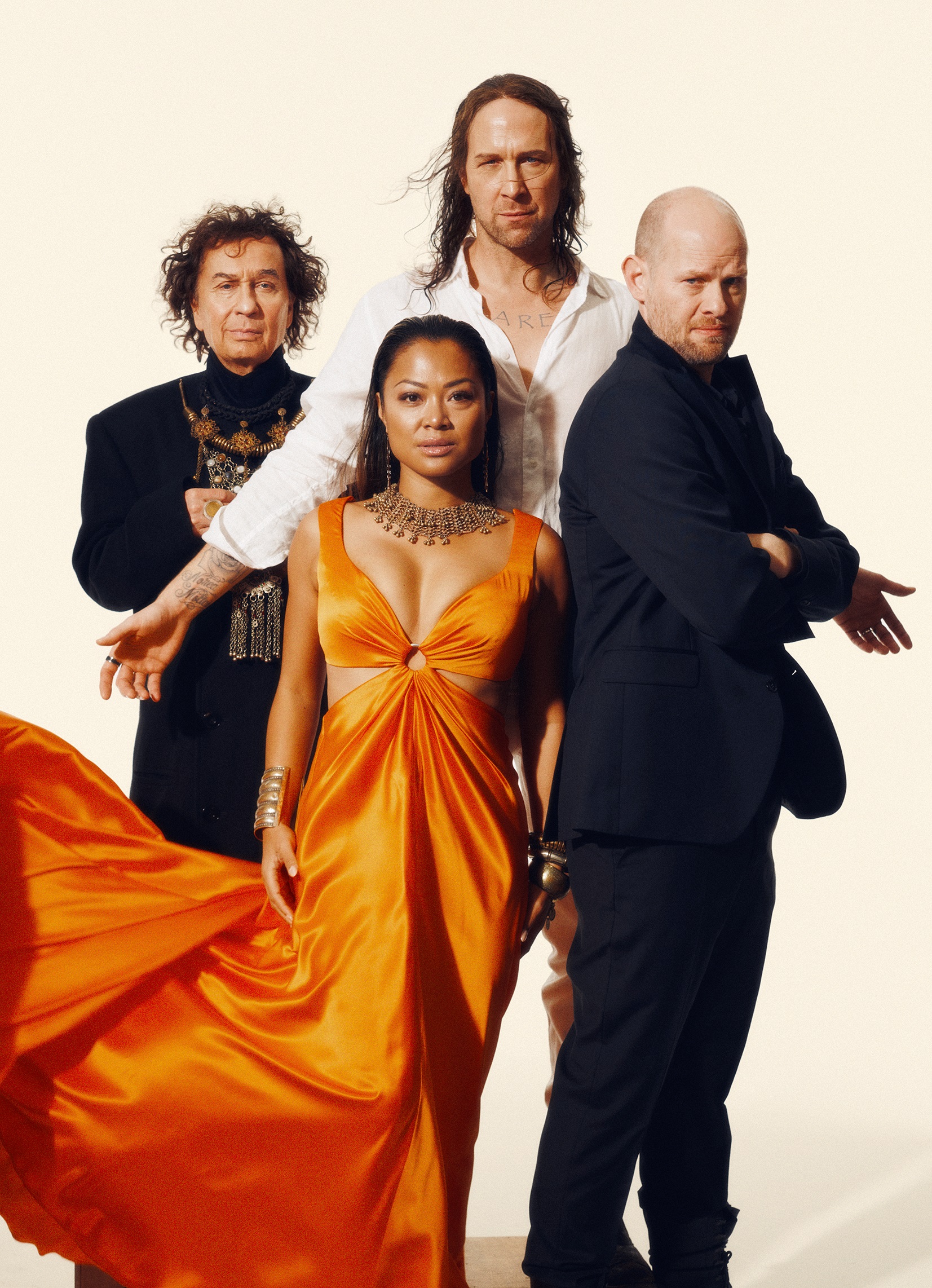 Fyra vuxna personer poserar: kvinna i orange klänning och tre män i mörka kläder bakom henne. En långhårig man i vit skjorta.