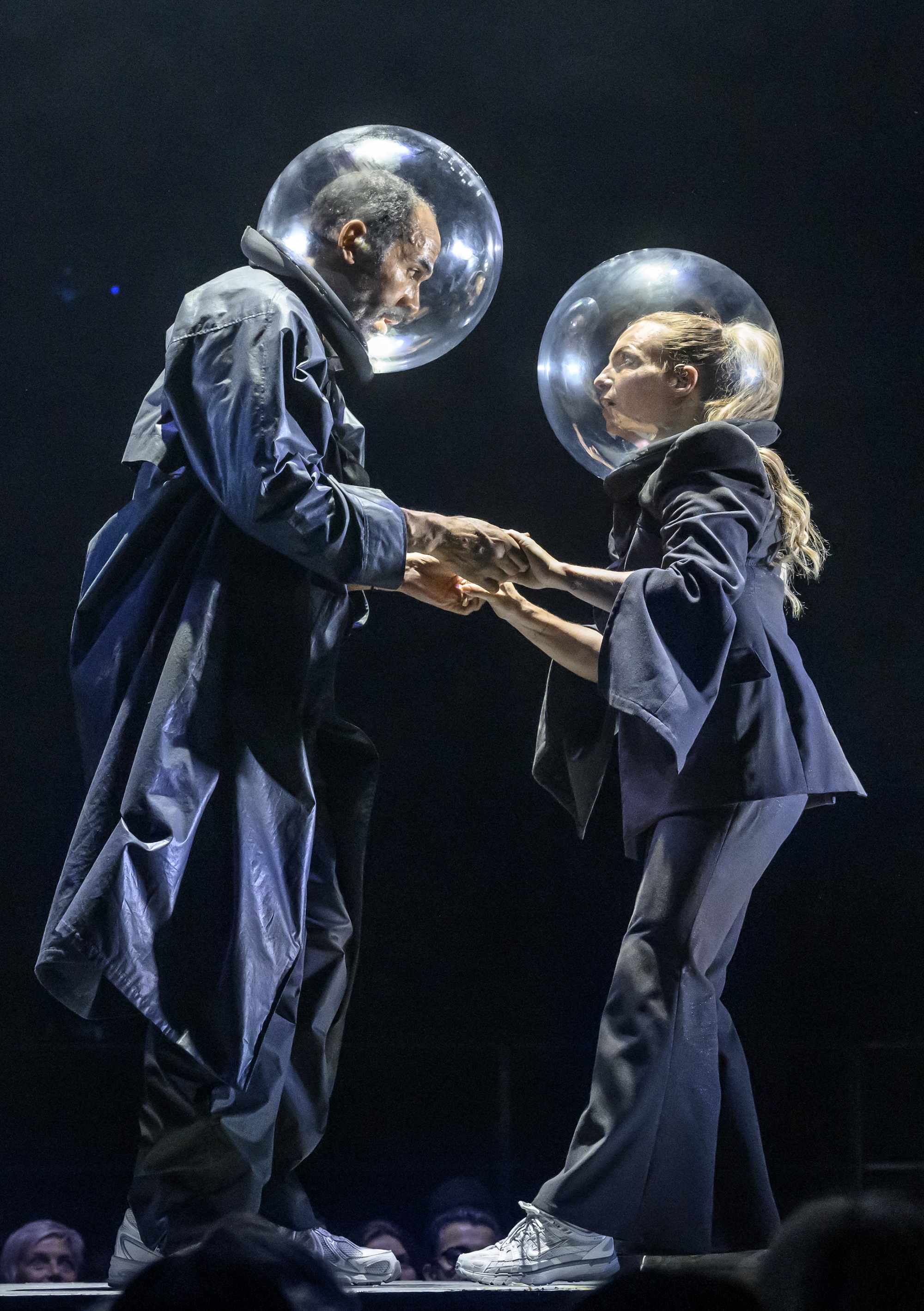 Man och kvinna i mörka kläder och stora genomskinliga hjälmar håller varandras händer på scen.