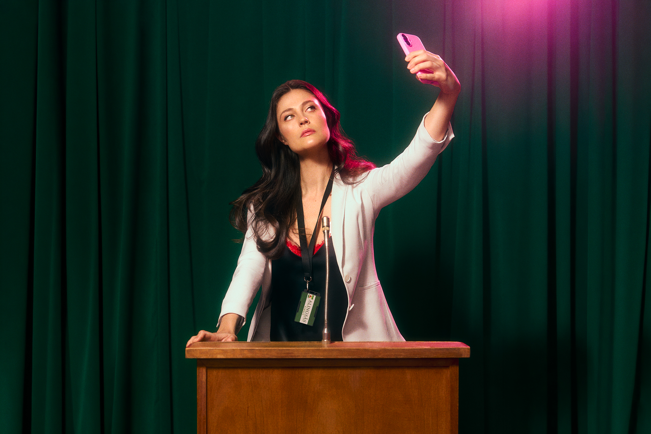 Kvinna vid en talarstol tar en selfie med mobilen, med namnbricka runt halsen och grön ridå i bakgrunden.