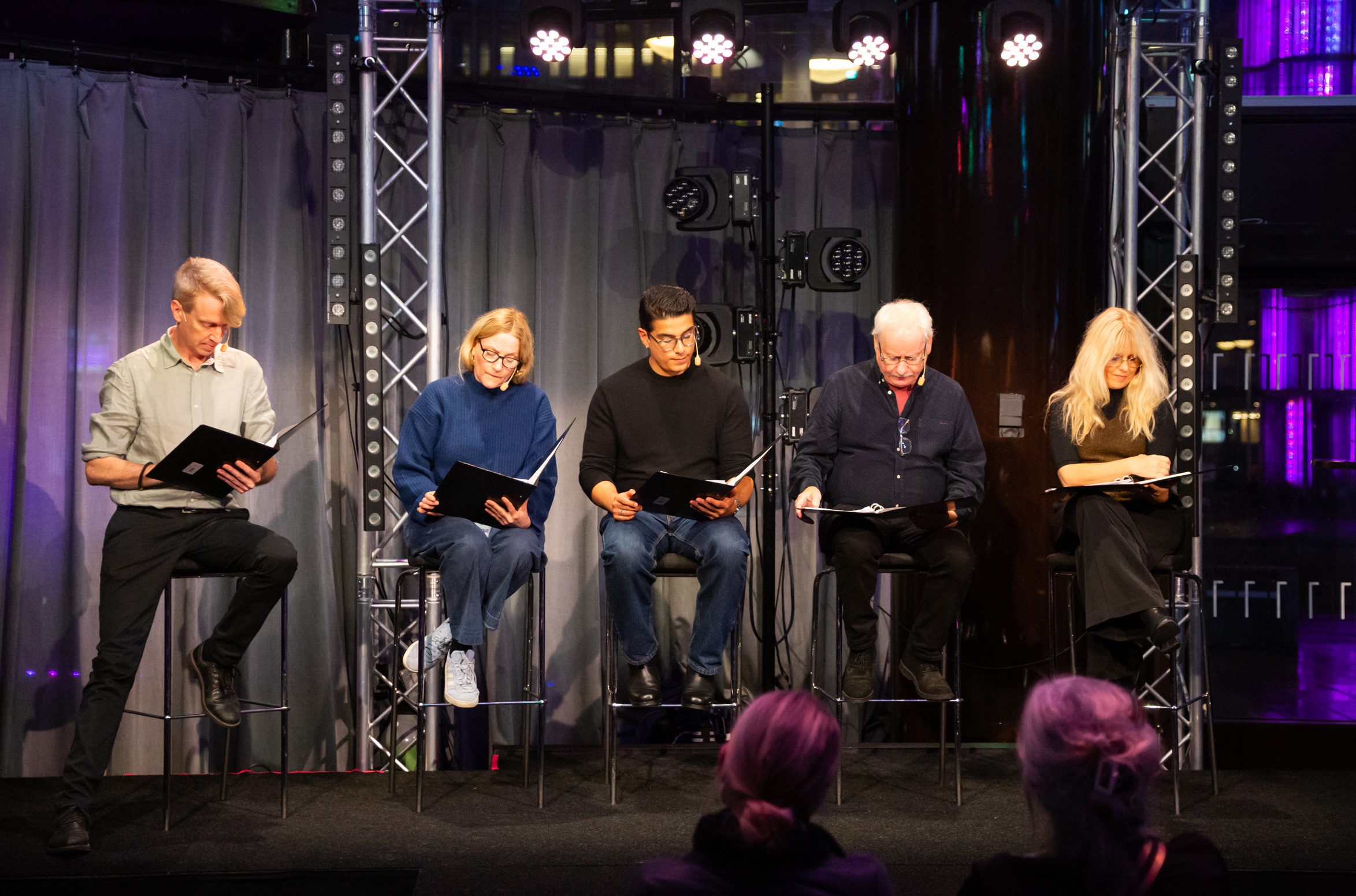 Sex personer, tre män och tre kvinnor i olika åldrar, sitter på pallar på scen och läser ur svarta manus.