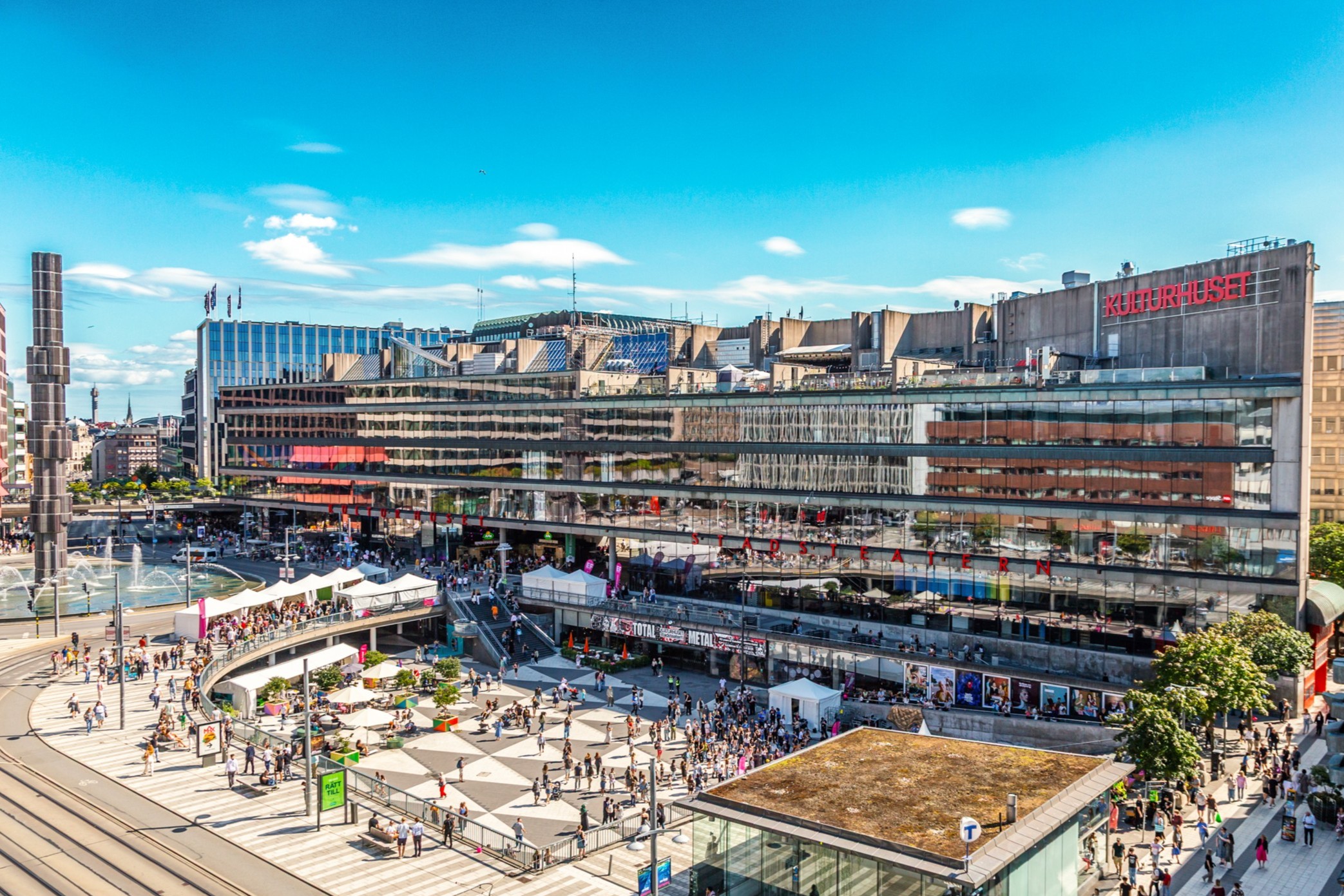 Flygvy över Kulturhuset vid Sergels torg i Stockholm, med många människor på torget och stadsliv i soligt väder.