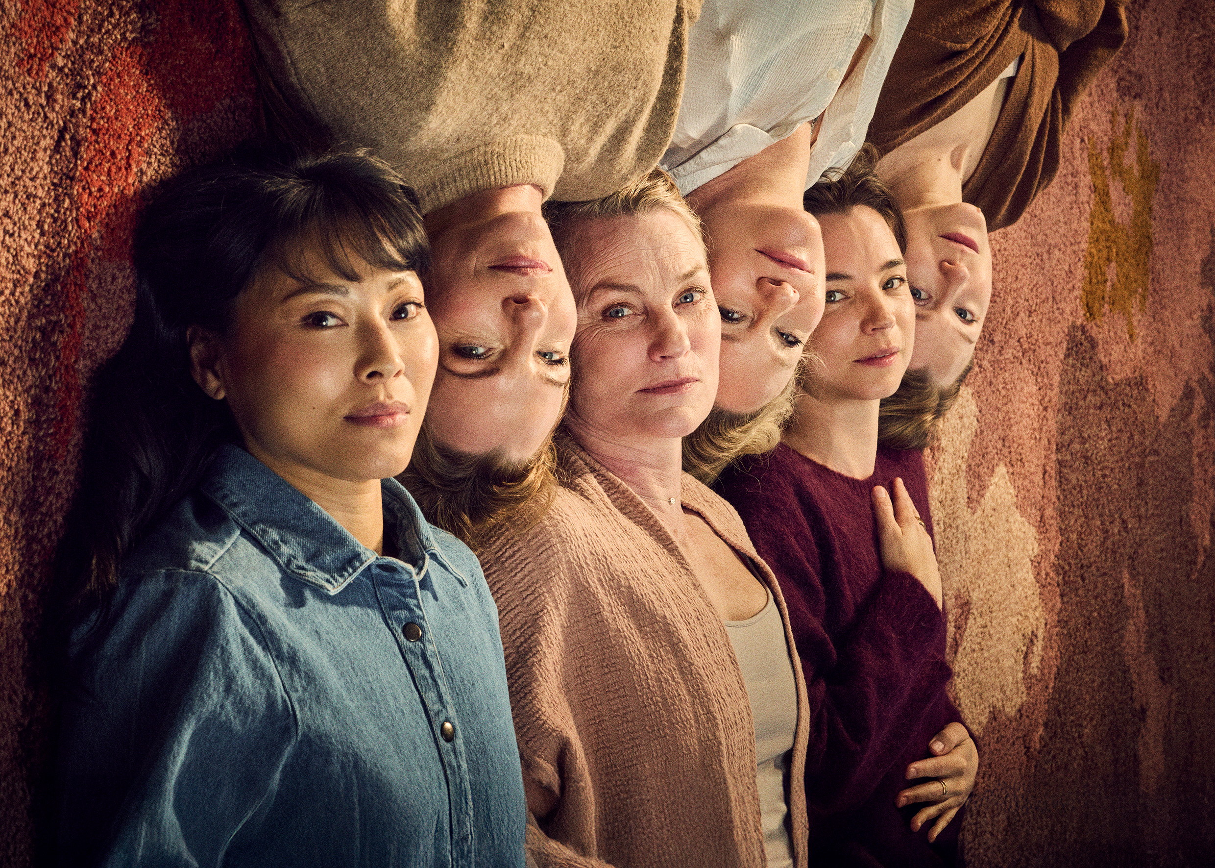 Sex kvinnor ligger tätt sida vid sida med huvuden mot varandra, sedda uppifrån mot en varmtonad bakgrund; de har olika åldrar och uttryck och blickar lugnt mot kameran.