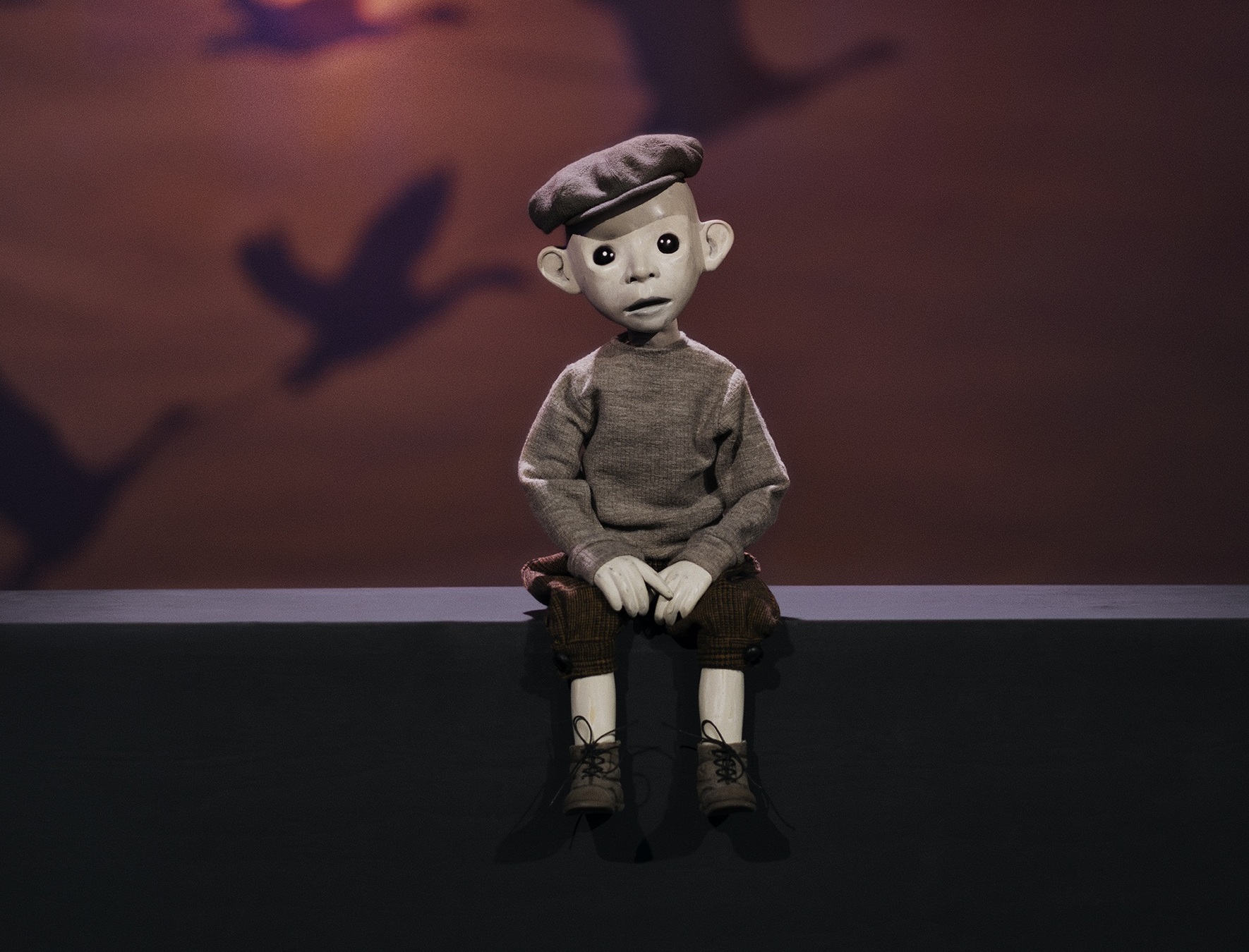 Docklik figur med basker sitter på en kant i dämpat ljus, med skuggor av flygande figurer projicerade på väggen bakom.